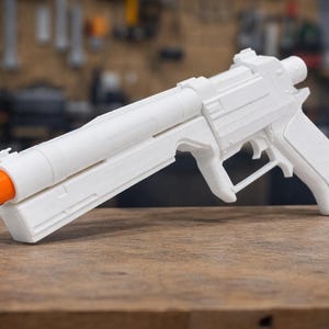 Könnte beinhalten: Eine weiße Spielzeugpistole mit orangefarbenem Lauf. Die Spielzeugpistole besteht aus Kunststoff und hat ein detailliertes Design, einschließlich Abzug, Griff und Visier. Sie liegt auf einer Holzoberfläche.