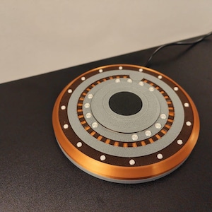 May include: A round, multi-layered wireless charger with a copper-colored outer ring, brown middle ring, and gray center. White dots and orange accents are visible. A black cable extends from the charger.