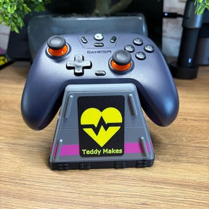 Peut inclure: Une manette Gamesir bleu foncé posée sur un support gris avec un cœur jaune et une bande rose. Le support porte le texte "Teddy Makes". La manette a des accents orange.