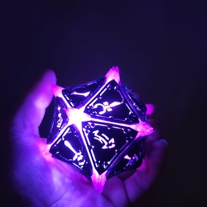 May include: A black and silver, multi-sided dice with glowing purple light emanating from the edges. The dice is being held in a hand.