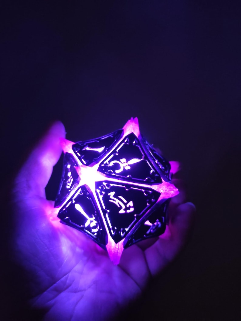 May include: A black and silver, multi-sided dice with glowing purple light emanating from the edges. The dice is being held in a hand.