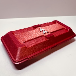 Könnte beinhalten: Ein rechteckiger, roter Behälter mit strukturierter Oberfläche und geometrischem Muster auf der Oberseite. Ein silbernes pfeilförmiges Design ist mittig auf dem Deckel angebracht. Der Behälter hat einen leicht erhöhten Rand.