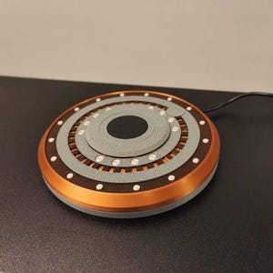 May include: A round, multi-layered wireless charger with a copper-colored outer ring, gray and brown inner rings, and a black center. Small white dots are evenly spaced around the charger. A black cord extends from the side.