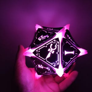 May include: A black and white illuminated dice with a sword symbol on one side. The dice is glowing pink.
