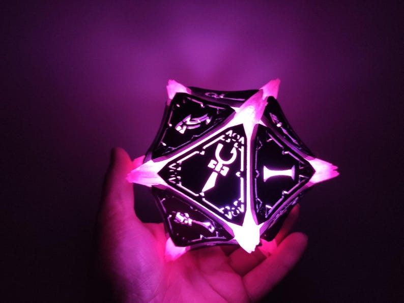 May include: A black and white illuminated dice with a sword symbol on one side. The dice is glowing pink.