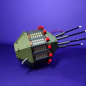 Könnte beinhalten: Ein geometrisches, olivgrünes Gerät mit roten Akzenten und mehreren schwarzen Stäben, die sich davon erstrecken. Das Gerät hat ein komplexes, geschichtetes Design mit goldenen und braunen Komponenten. Der Hintergrund ist ein tiefes Lila.