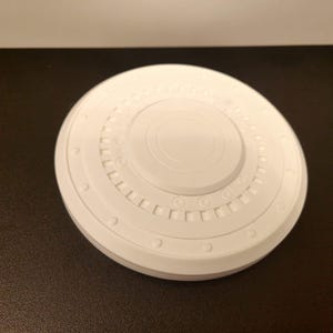 May include: A white, circular object with a detailed, layered design. The object has a central raised circle and multiple concentric rings with small, evenly spaced details. The overall shape is reminiscent of a spacecraft component.