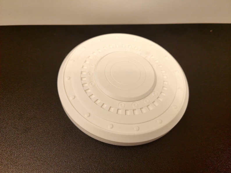 May include: A white, circular object with a detailed, layered design. The object has a central raised circle and multiple concentric rings with small, evenly spaced details. The overall shape is reminiscent of a spacecraft component.