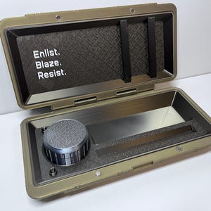 Könnte beinhalten: Ein offener olivgrüner Koffer mit strukturiertem Innenraum und einem silberfarbenen Zifferblatt. Der Koffer trägt die Aufschrift "Enlist. Blaze. Resist.". Der Koffer ist für kleine Gegenstände konzipiert.