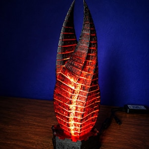 May include: A vertical, double-pointed sculpture featuring a dark, textured exterior covered in etched symbols. The object emits a bright, internal red glow from between its segments and rests on a dark, hexagonal base with a power cord attached.