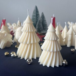 Puede incluir: Una colección de velas blancas con forma de árbol de Navidad, algunas con un diseño texturizado y plisado, y otras con un acabado liso. Las velas están dispuestas sobre una superficie azul oscuro con pequeñas campanas plateadas esparcidas a su alrededor.