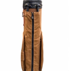 May include: A brown golf bag with a black zip and detailing. The bag features a leather top and base, with a front and side pocket. Designed to carry golf clubs and accessories.