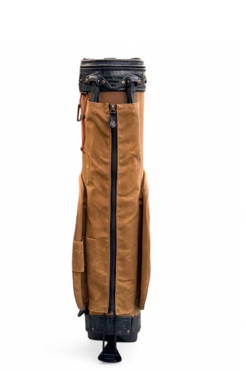 May include: A brown golf bag with a black zip and detailing. The bag features a leather top and base, with a front and side pocket. Designed to carry golf clubs and accessories.