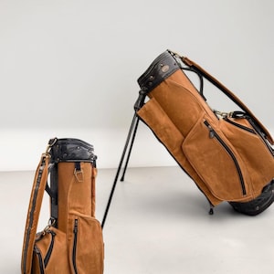 May include: Two brown golf bags with black detailing. One bag stands upright, the other is on its stand. The bags feature multiple zipped pockets and a black top with a handle. The bags are made of a suede-like material.