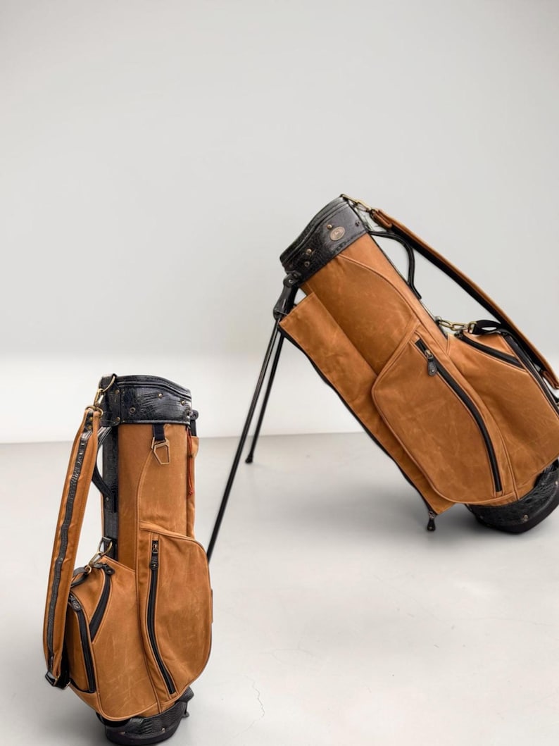 May include: Two brown golf bags with black detailing. One bag stands upright, the other is on its stand. The bags feature multiple zipped pockets and a black top with a handle. The bags are made of a suede-like material.
