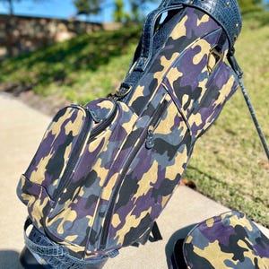 May include: A golf bag featuring a camouflage design in purple, beige, and grey. The bag has dark blue faux-crocodile leather trim and a handle. The bag is on a concrete surface with a matching cover.