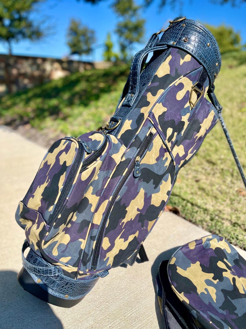 May include: A golf bag featuring a camouflage design in purple, beige, and grey. The bag has dark blue faux-crocodile leather trim and a handle. The bag is on a concrete surface with a matching cover.