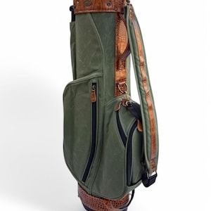May include: Green golf bag with brown leather detailing. The bag has multiple zipped pockets, a padded shoulder strap, and a textured leather base and top. The bag has a classic design.