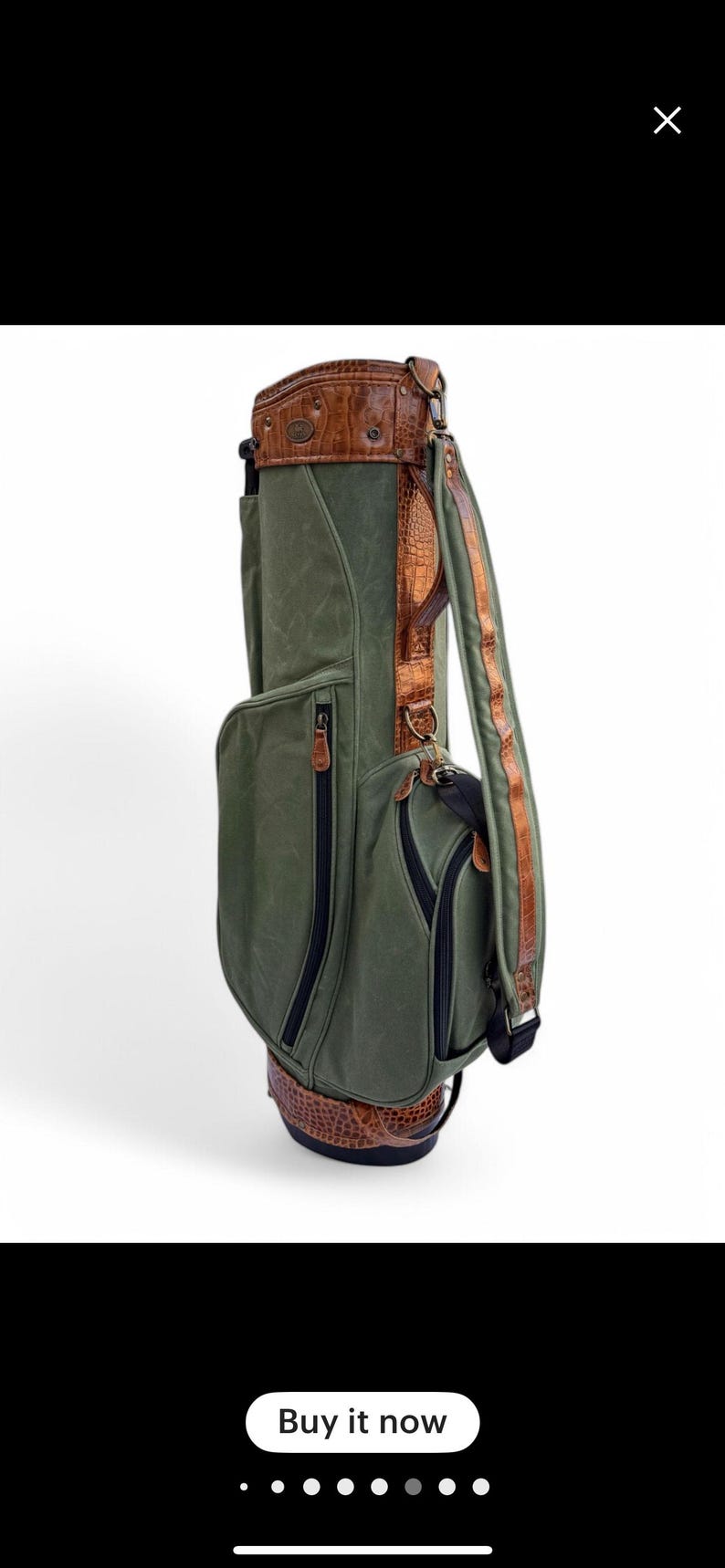 May include: Green golf bag with brown leather detailing. The bag has multiple zipped pockets, a padded shoulder strap, and a textured leather base and top. The bag has a classic design.