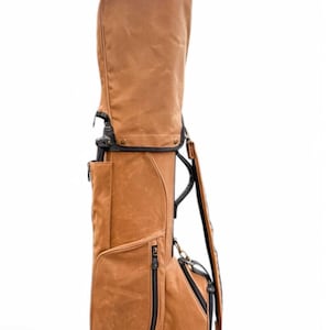 May include: A brown golf bag with a structured design. The bag features multiple zippered pockets, a shoulder strap, and a top cover. The bag is made of a textured material and has black accents. The bag is designed to hold golf clubs.
