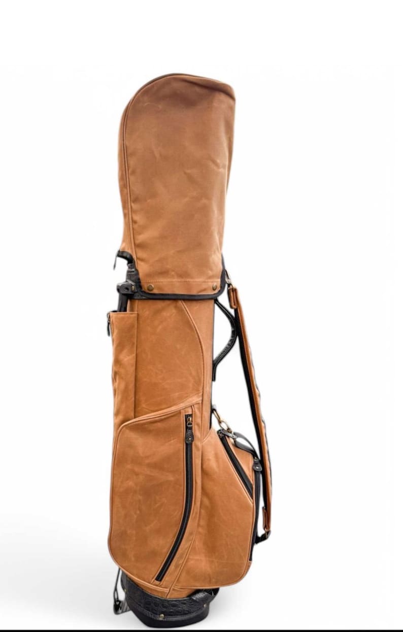 May include: A brown golf bag with a structured design. The bag features multiple zippered pockets, a shoulder strap, and a top cover. The bag is made of a textured material and has black accents. The bag is designed to hold golf clubs.