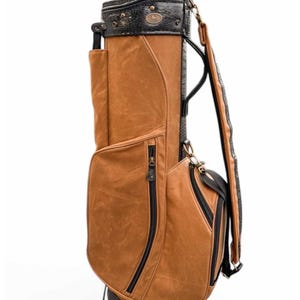 May include: A brown and black golf bag with multiple pockets and a shoulder strap. The bag has a textured leather appearance with black zips and trim. The top section is black with a decorative emblem.