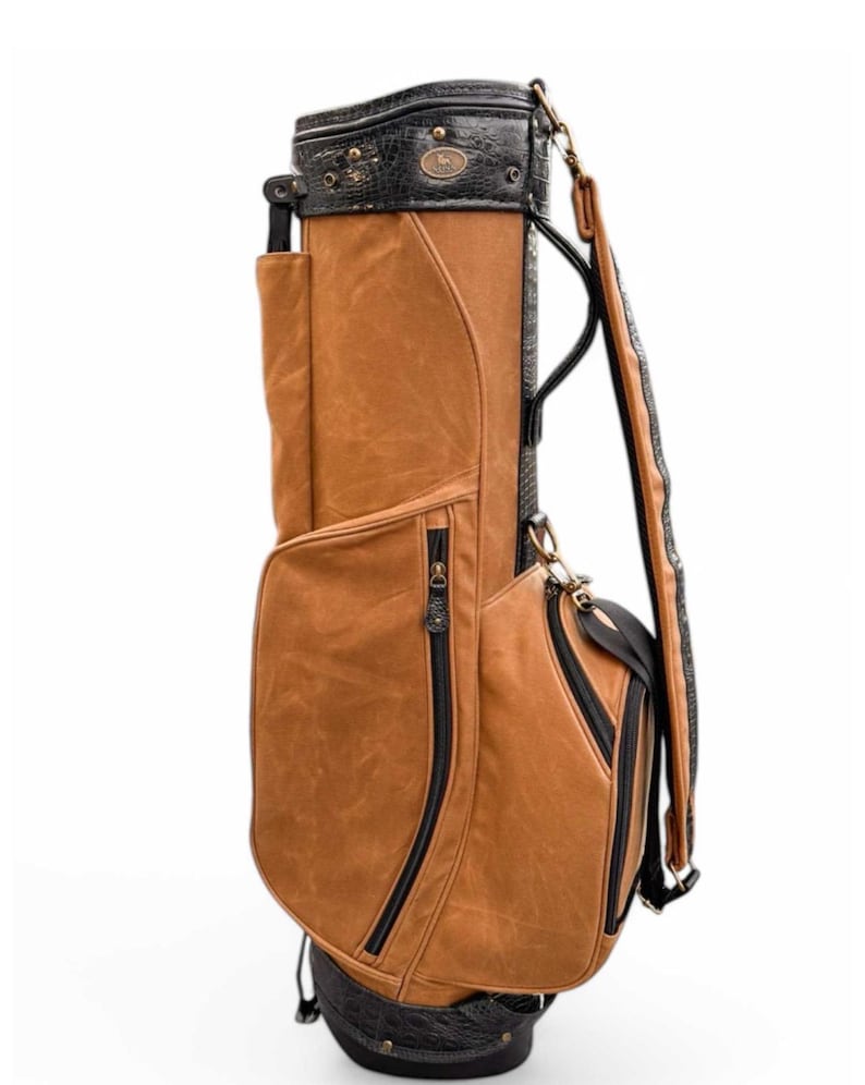 May include: A brown and black golf bag with multiple pockets and a shoulder strap. The bag has a textured leather appearance with black zips and trim. The top section is black with a decorative emblem.