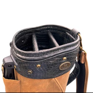 May include: A golf bag with a black, textured top and a tan body. The top has multiple compartments and is secured with brass-coloured rivets. A shoulder strap and a small logo are visible.