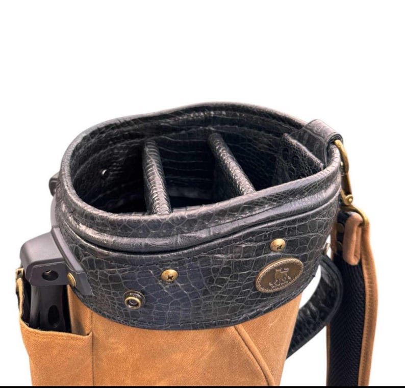 May include: A golf bag with a black, textured top and a tan body. The top has multiple compartments and is secured with brass-coloured rivets. A shoulder strap and a small logo are visible.
