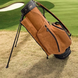 May include: A brown golf bag with black detailing and a stand. The bag is made of a textured material and has multiple pockets with black zips. The top of the bag is black with a decorative pattern. The bag is on grass.