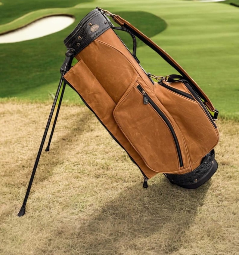 May include: A brown golf bag with black detailing and a stand. The bag is made of a textured material and has multiple pockets with black zips. The top of the bag is black with a decorative pattern. The bag is on grass.