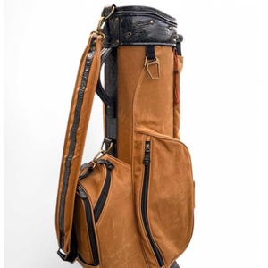 May include: A brown golf bag with black detailing. The bag has a textured, leather-like finish, black zips, and a black top with a crocodile-style pattern. A shoulder strap is included for carrying.