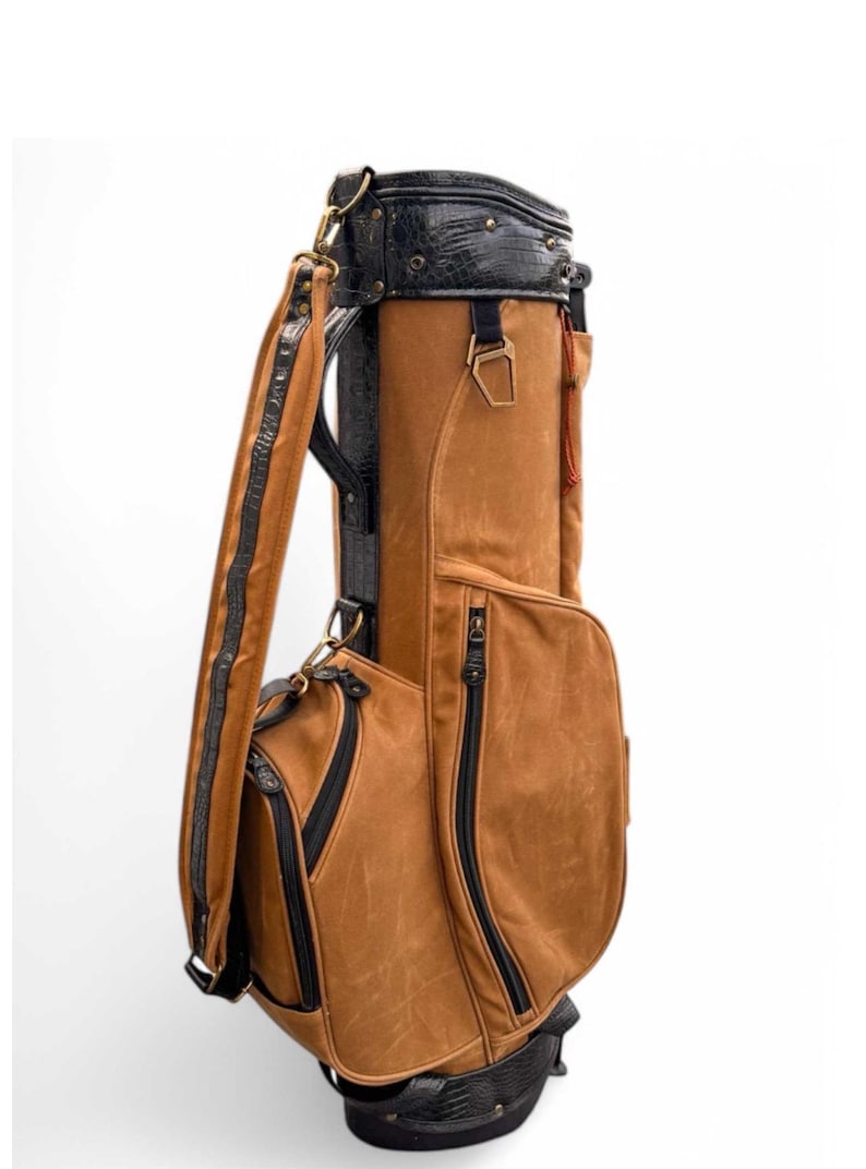 May include: A brown golf bag with black detailing. The bag has a textured, leather-like finish, black zips, and a black top with a crocodile-style pattern. A shoulder strap is included for carrying.