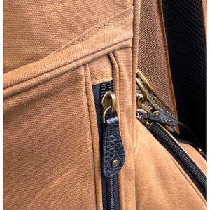 May include: Close-up of a brown canvas backpack with a black zip and a leather zip pull. The zip pull is dark blue with a textured pattern and a brass-coloured metal zip pull.