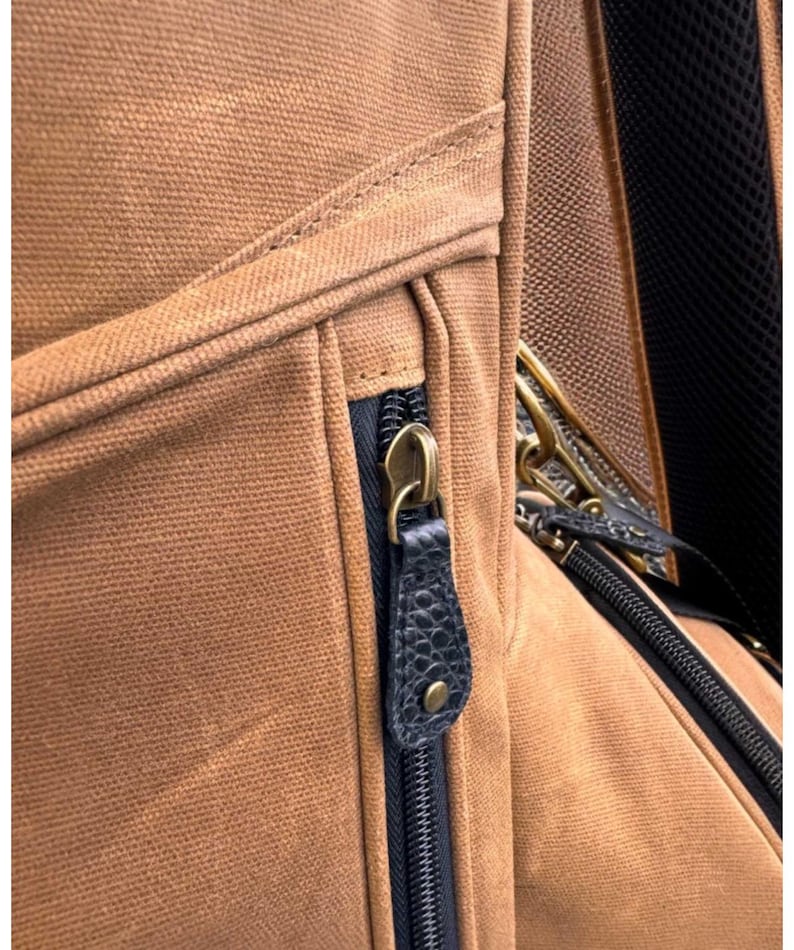 May include: Close-up of a brown canvas backpack with a black zip and a leather zip pull. The zip pull is dark blue with a textured pattern and a brass-coloured metal zip pull.