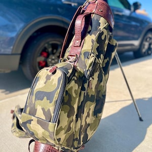 May include: Camouflage golf bag with brown leather detailing. The bag displays a camouflage pattern in green, brown, and black. The top and base are trimmed with brown leather, and it has a matching brown leather handle.