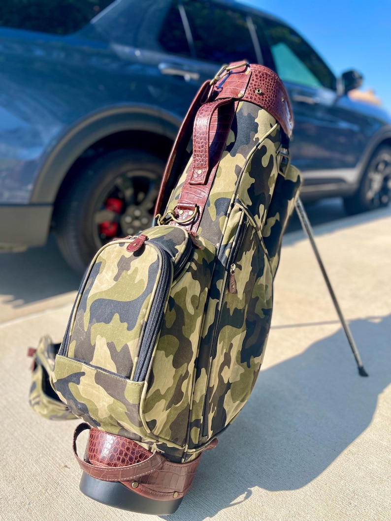 May include: Camouflage golf bag with brown leather detailing. The bag displays a camouflage pattern in green, brown, and black. The top and base are trimmed with brown leather, and it has a matching brown leather handle.