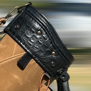 May include: Black leather bag with a crocodile skin texture on the top flap. The bag has brass-coloured hardware and a brown canvas body. A brown leather cord hangs from the bag.