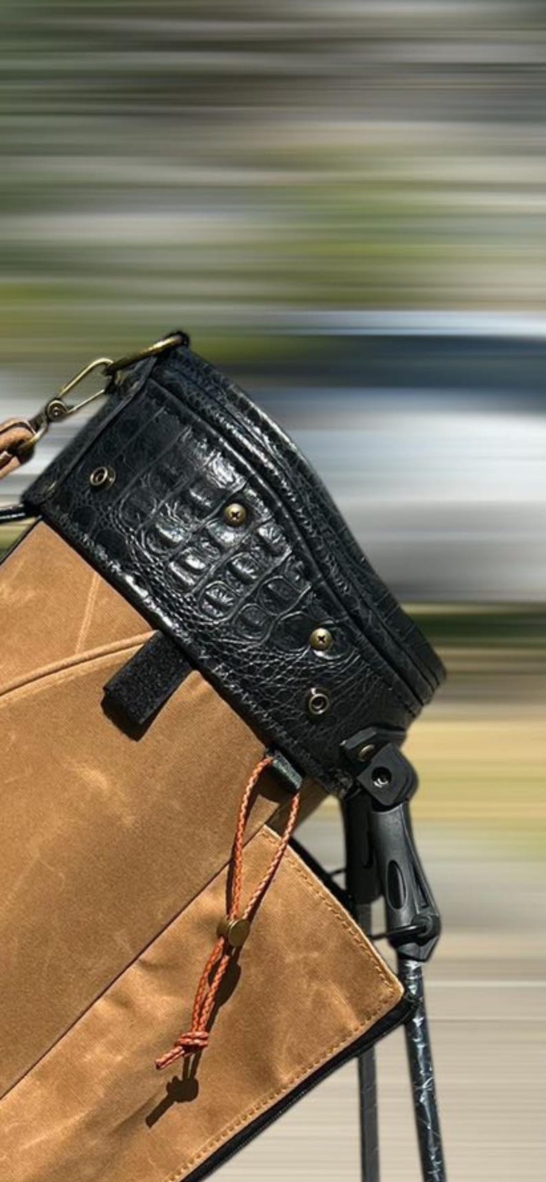 May include: Black leather bag with a crocodile skin texture on the top flap. The bag has brass-coloured hardware and a brown canvas body. A brown leather cord hangs from the bag.