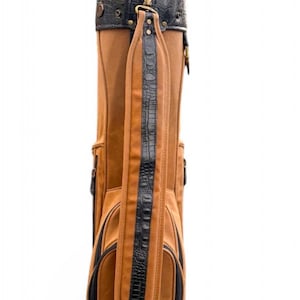 May include: A brown golf bag with black detailing. The bag combines brown leather with black faux crocodile leather on the straps and top. It has a shoulder strap and several pockets. The bag is designed for carrying golf clubs.