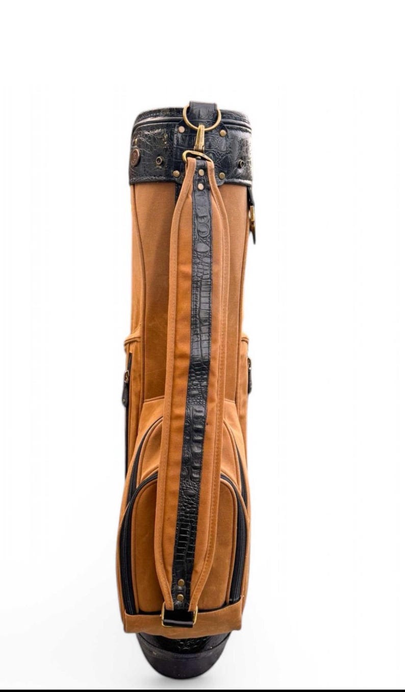 May include: A brown golf bag with black detailing. The bag combines brown leather with black faux crocodile leather on the straps and top. It has a shoulder strap and several pockets. The bag is designed for carrying golf clubs.