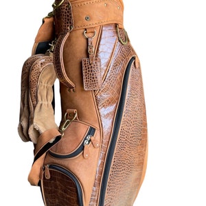 May include: A brown golf bag with a combination of smooth and textured leather. The bag features a crocodile-like embossed pattern on the side panels and pockets, with a suede-like finish on the top and handle. It has multiple pockets, zippers, and a shoulder strap.