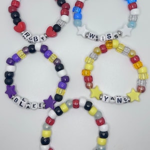 May include: Five colorful beaded bracelets with different names spelled out on them. The bracelets are made with white, red, blue, yellow, purple, black, and clear beads. The names on the bracelets are "Ruby", "Weiss", "Blake", "Yang", and "RWBY".