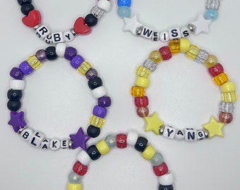 May include: Five colorful beaded bracelets with different names spelled out on them. The bracelets are made with white, red, blue, yellow, purple, black, and clear beads. The names on the bracelets are "Ruby", "Weiss", "Blake", "Yang", and "RWBY".