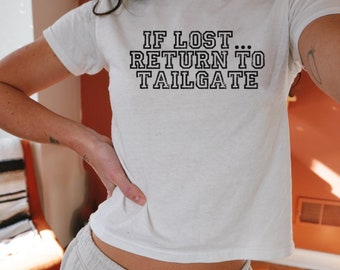 May include: White cropped t-shirt with the text "IF LOST... RETURN TO TAILGATE" in black. The model is wearing the shirt with white sweatpants and the text "WHITE - XS" is visible at the bottom of the image.
