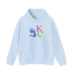 May include: A light blue hoodie with a front pocket and a drawstring hood. The front features a graphic of a blue hand embracing a smaller hand, with the letters "KC" in pink and green.