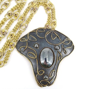 May include: A vintage necklace featuring a large, abstract pendant with a dark gray center and brass-colored accents. The pendant has an oval-shaped stone and is suspended from a brass chain. The necklace is set against a white background.