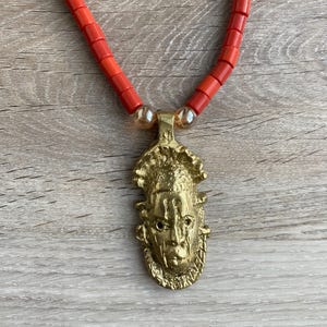 Puede incluir: Un collar de color dorado con un colgante que presenta un rostro humano estilizado. El collar está hecho de cuentas rojas.