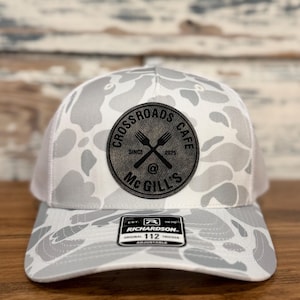 May include: A gray and white camouflage trucker hat with a white mesh back. The front of the hat features a circular patch with the text "Crossroads Cafe Since 2025 @ McGill's" and crossed forks. The hat is adjustable.