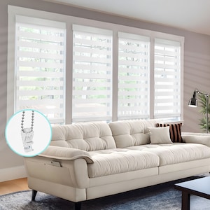 May include: White zebra blinds with a chain and bead pull. The blinds are installed in a living room with a white couch and a black lamp.